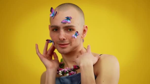 Porträtt av glada manliga kaukasiska queer med fjärilar i ansiktet i klänning och blommigt halsband poserar på gul bakgrund. Självsäker stolt man tittar på kameran leende. — Stockvideo