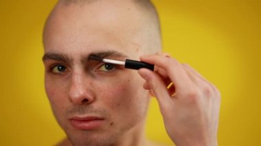 Kameraya bakarak rimel uygulayan kendinden emin erkek transseksüelin yakın görüntüsü. Sarı arka planda makyaj yaparken poz veren genç beyaz erkek portresi. Kadınsı erkek..