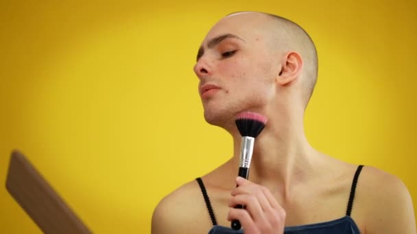 Αρσενικό queer εφαρμογή πούδρα προσώπου και κοιτάζοντας κάμερα με αυτοπεποίθηση έκφραση προσώπου. Πορτρέτο του Καυκάσου νεαρού άνδρα που ποζάρει στο κίτρινο φόντο μακιγιάροντας. — Αρχείο Βίντεο