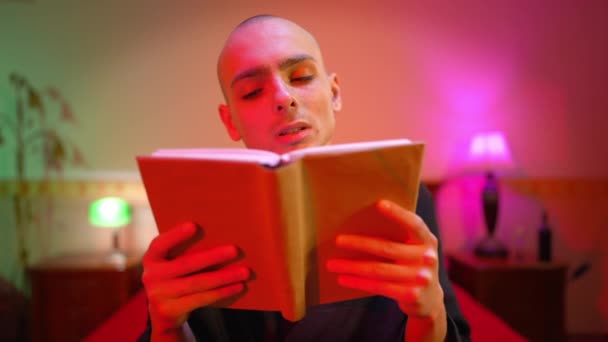 Portret van een jonge man in een jurk die het slotboek leest en naar de camera kijkt met droevige gezichtsuitdrukking. Vooraanzicht van mannelijke blanke homo poseren in de slaapkamer in rood licht. — Stockvideo