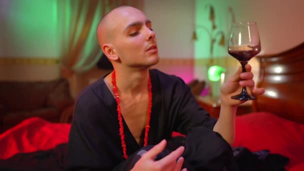 Confident male queer shaking red wine in glass looking at camera sitting in bedroom with red light. Portrait of feminine Caucasian man in dress posing with alcohol at home indoors. — Stock Video