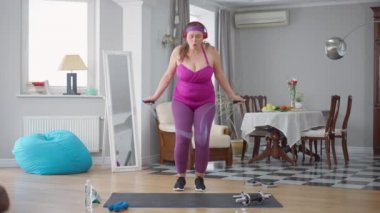 Evinde ip atlayıp zıplayan obez beyaz kadının ön manzara portresi. Yüksek kilolu kadın iç mekanda antrenman yapıyor. Kilo kaybı ve vücut pozitifliği.