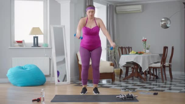 Επίμονη σοβαρά κίνητρα παχύσαρκη γυναίκα ανύψωσης αλτήρες κατάρτισης στο σπίτι σε εσωτερικούς χώρους. Ευρεία μπροστινή όψη υπέρβαρης Καυκάσιας κυρίας που γυμνάζεται στην άρση βαρών. Αθλητισμός και άσκηση. — Αρχείο Βίντεο