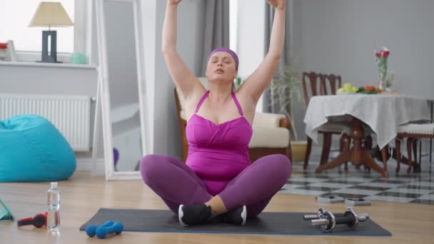 Портрет толстой белой молодой женщины в позе лотоса, делающей дыхательные упражнения. Вид спереди широкий снимок тренировки тысячелетия ожирения, медитирующей дома, засыпающей. Концепция йоги и тренировок. — стоковое видео