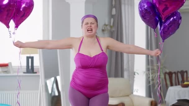 Μέτριο πλάνο της αστεία παχύσαρκη γυναίκα αύξηση μπαλόνια που μιμούνται την άρση βάρους κοιτάζοντας την κάμερα. Πορτρέτο της χαλαρής απρόσεκτη χαρούμενη υπέρβαρη κυρία θέτει σε εσωτερικούς χώρους στο σπίτι. Σωματικό και χιούμορ. — Αρχείο Βίντεο