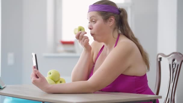 Vista lateral da jovem caucasiana de tamanho grande que come maçã de vitamina orgânica saudável olhando para a tela do smartphone. Retrato de inspiração motivado dieta senhora obesa para perda de peso. — Vídeo de Stock