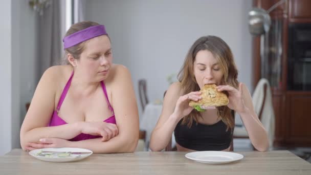 Jaloers dikke vrouw kijkt naar slanke blanke vriend eten sandwich. Vooraanzicht van slanke en zwaarlijvige vrouwen die aan tafel zitten met gezond en ongezond voedsel. Obesitas en schoonheid concept. — Stockvideo