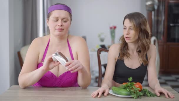 Retrato de mujer caucásica obesa comiendo chocolate delicioso dulce como delgada mujer arrogante masticando hojas de ensalada verde. Feliz sobrepeso millennial con amigo delgado en el interior. Cuerpo positivo y elección. — Vídeo de stock