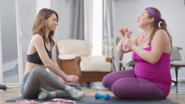 Side view portræt af smilende positiv smuk slank kvinde giver forfriskende vand til ophidset overvægtige ven taler hvile efter hjemmetræning. Kaukasiske bofæller på workout pause indendørs. – Stock-video