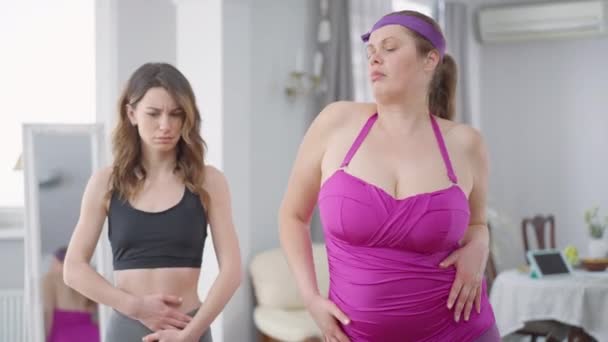 Selvsikker overvægtig kvinde og usikker slank årtusinde stående indendørs poserende. Portræt af kaukasiske overvægtige dame og slank værelseskammerat derhjemme. Tillid og krop positiv. – Stock-video