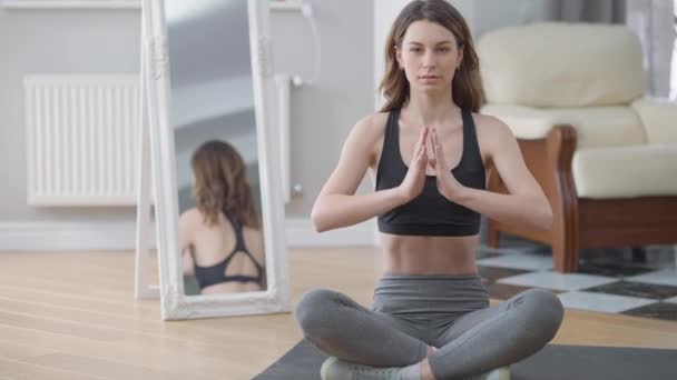 Mujer caucásica hermosa y segura sentada en pose de loto haciendo ejercicio en casa. Amplia vista frontal de la hermosa morena deportista yogui de entrenamiento en interiores. Tranquilidad y yoga. — Vídeos de Stock