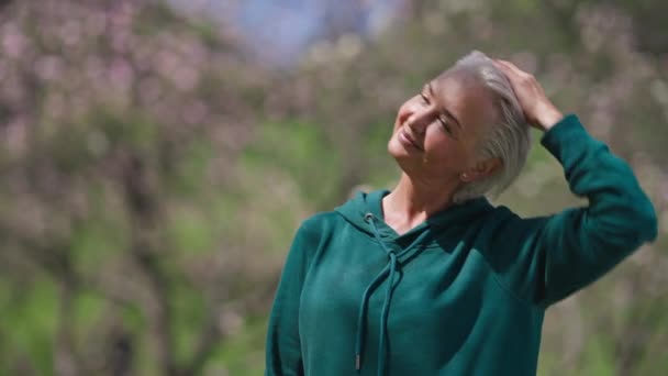 Vidám idős nő nyújtózkodó nyaki izmokat edz a napos nyári parkban. Portré pozitív gyönyörű vékony mosolygó kaukázusi nyugdíjas gyakorló szabadban. Sport és életmód koncepció. — Stock videók