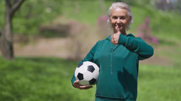 Portrait de femme âgée caucasienne joyeuse tenant ballon de football montrant pouce vers le haut en souriant. Joyeux retraité en forme posant dans le parc d'été de l'exercice à l'extérieur. Vieillissement et mode de vie sain. — Video