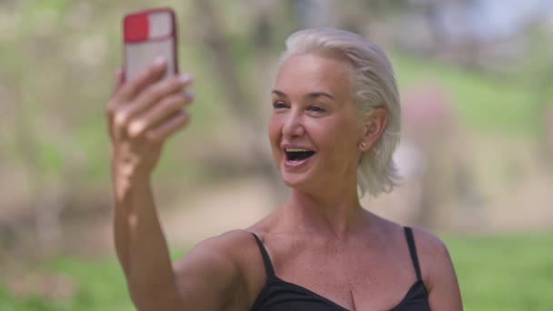 Alegre mujer mayor de confianza hablando utilizando el chat de vídeo en el teléfono inteligente de pie en el soleado parque de verano. Retrato de la deportista caucásica feliz jubilada charlando en el entrenamiento al aire libre. — Vídeos de Stock