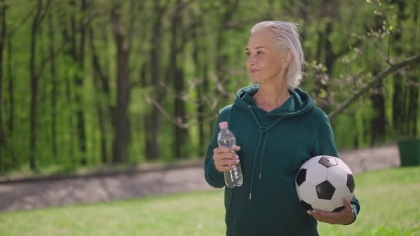 Zoom caméra en direct pour sourire satisfait sportif caucasien senior debout avec ballon de football et bouteille d'eau dans le parc d'été ensoleillé. Joyeux entraînement pour retraités en plein air. Mouvement lent. — Video