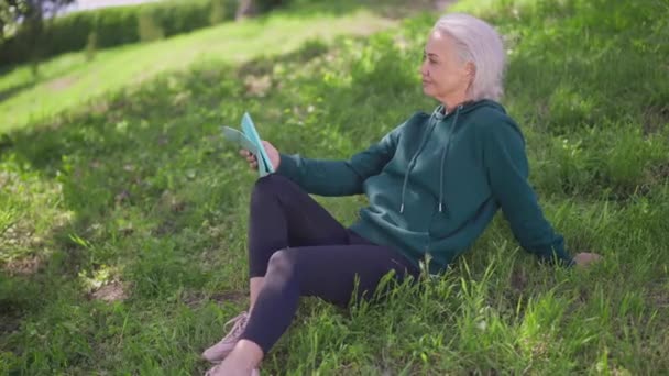 Oldalnézet mosolygós idős nő ül a zöld nyári réten tabletta gondolkodás. Magabiztos kaukázusi nyugdíjas élvezi a közösségi média görgetés a szabadban a napsütéses napon. Szabadidős koncepció. — Stock videók