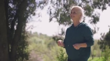 Güneşli yaz parkında ağır çekimde koşan mutlu yaşlı kadın gülümsüyor. Kendine güvenen beyaz emeklinin portresi gün ışığında dışarıda koşmaktan zevk alıyor. Spor ve eğlence konsepti.