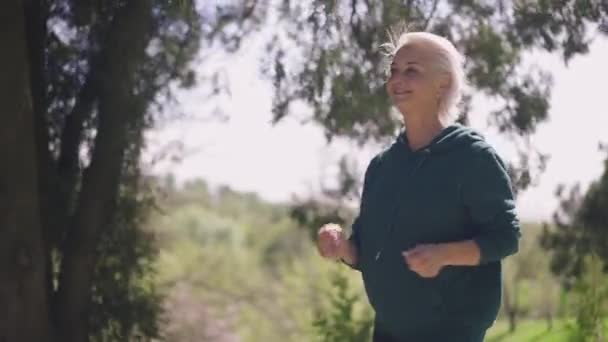 Szczęśliwa seniorka biegająca w zwolnionym tempie w słonecznym letnim parku uśmiechnięta. Portret pewnego siebie białego emeryta cieszącego się joggingiem na świeżym powietrzu w świetle słonecznym. Sport i koncepcja radości. — Wideo stockowe