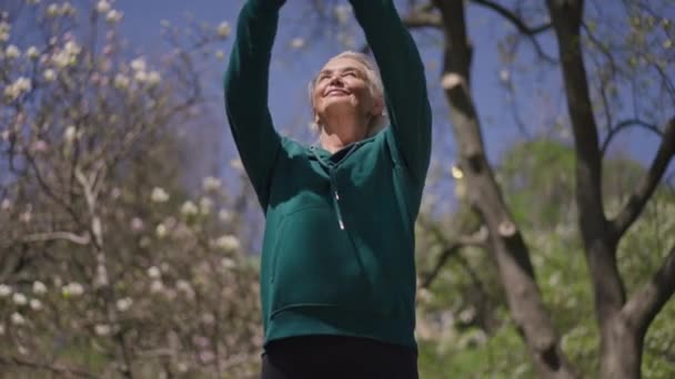 Elégedett idős, sportos nő felemeli a kezét mosolyogva áll a nyári parkban virágzó fák a háttérben. A boldog fehér nyugdíjas kiképzés portréja a szabadban. Sport és boldogság. — Stock videók