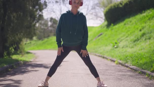 Amplia toma de confiada deportista senior calentándose en cámara lenta mirando a la cámara. Retrato de mujer caucásica en auriculares estirando las piernas a la luz del sol al aire libre preparándose para trotar. — Vídeo de stock