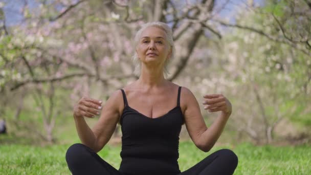 Framifrån av säker passform senior idrottskvinna sitter i lotus utgör meditera i vår sommarpark. Porträtt av smal kaukasisk kvinnlig pensionär som utövar yoga på solig dag utomhus. Långsamma rörelser. — Stockvideo