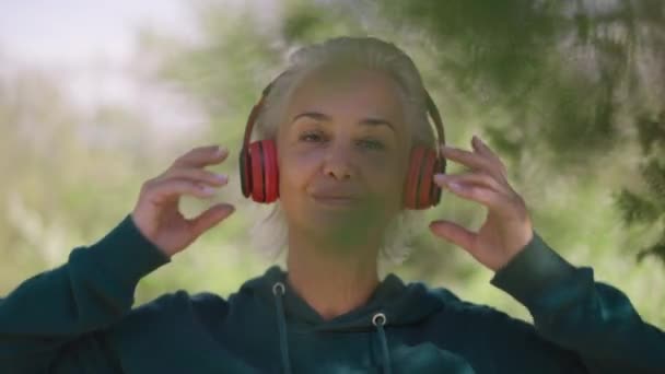 Close-up portret van een zelfverzekerde senior fitte sportvrouw die een koptelefoon opzet die naar de camera kijkt en traint in slow motion. Prachtige blanke gepensioneerde training in het voorjaar zonnig park buiten. — Stockvideo