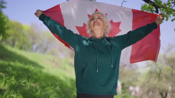 A boldog nyugdíjas portréja, amint kanadai zászlót lobogtatva, kinyújtott kezekkel néz félre a napsütéses napon. Mosolygó fehér idős nő csomagolva nemzeti szimbólum lassított felvételen. — Stock videók