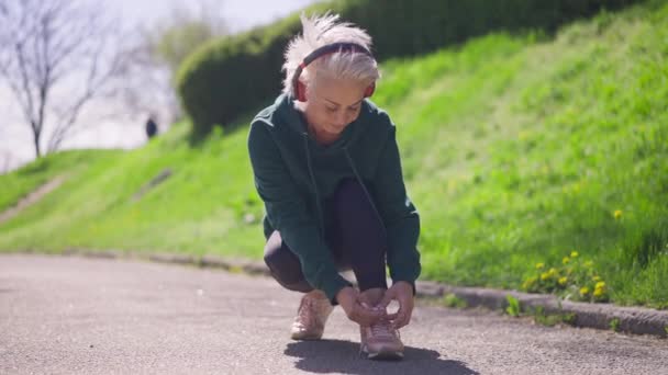 Biała, starsza sportowiec zawiązuje sznurowadła i ucieka w zwolnionym tempie. Pozytywny emeryt pewny siebie w słuchawki jogging w słonecznym parku na świeżym powietrzu. Styl życia i koncepcja sportu. — Wideo stockowe