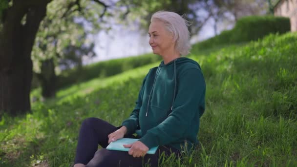 Souriant heureuse femme caucasienne âgée montrant pouce vers le haut assis sur la pelouse d'été de printemps avec tablette. Portrait de retraité mince confiant profitant d'une journée ensoleillée se reposant à l'extérieur. Mouvement lent. — Video