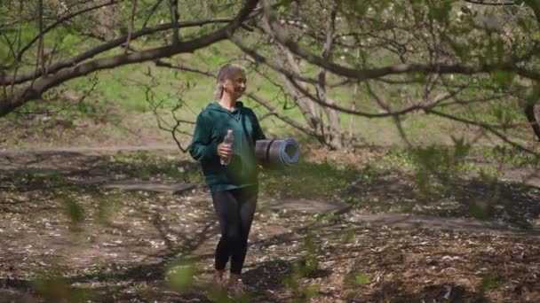 Amplio tiro de sonriente confiada deportista senior caminando en cámara lenta en el parque de verano con colchoneta de ejercicio y botella de agua. Feliz Caucásico jubilado entrenamiento al aire libre en día soleado. — Vídeos de Stock