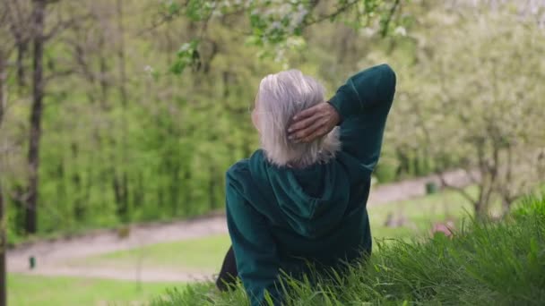 Bakåt syn på avslappnad gråhårig senior kvinna sitter på grön gräsmatta njuter solig dag på våren sommarpark. Glad bekymmerslös kaukasisk pensionär vilar utomhus. Långsamma rörelser. — Stockvideo