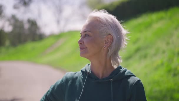 Magabiztos, ősz hajú, barna szemű, mosolygó sportnő, aki a kamerát nézi a reggeli edzés alatt a parkban. Portré boldog boldog boldog kaukázusi nyugdíjas gyakorló napfény szabadban. — Stock videók