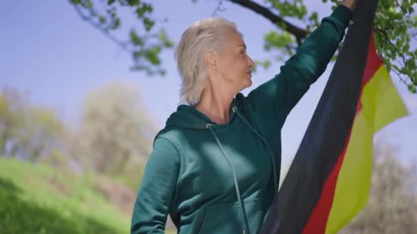 Drapeau allemand flottant dans les mains des femmes comme souriant fière femme âgée confiante détournant les yeux. Portrait de belle retraitée debout dans le parc avec le symbole national souriant. Concept de patriotisme. — Video