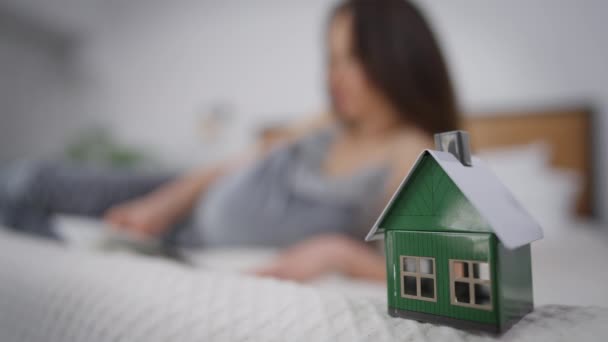 Close-up de casa de brinquedo na cama com mulher branca grávida borrada lendo livro deitado no fundo. Jovem morena expectante desfrutando de hobby dentro de casa no quarto em novo apartamento. — Vídeo de Stock