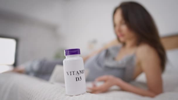 Vitamine D3 fles op bed met wazig zwangere vrouw lezen boek liggend op de achtergrond. Brunette Kaukasische jonge verwachting genieten van hobby thuis binnen met pillen aan de voorkant. Gezondheidszorg en zwangerschap. — Stockvideo