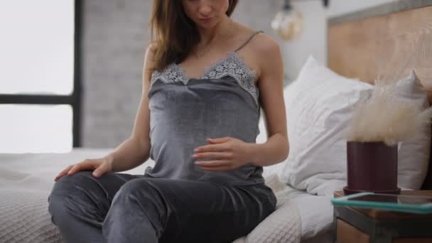 Ugenkendelig slank kaukasisk gravid kvinde kærtegner mave sidder på sengen derhjemme. Unge forventningsfulde i pyjamas strøg mave om morgenen indendørs i slowmotion. Graviditet koncept. – Stock-video
