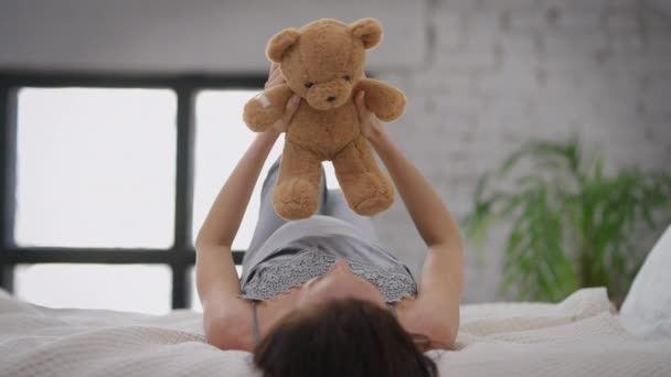 Teddy beer in handen van jonge zwangere blanke vrouw die op bed ligt te spelen met speelgoed. Brunette ontspannen vrolijke verwachting plezier hebben thuis binnen. Langzame beweging. — Stockvideo