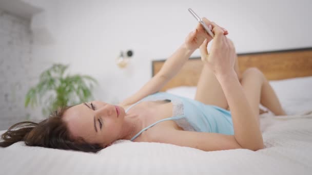 Νεαρή λεπτή μελαχρινή λευκή γυναίκα με μπλε πιτζάμες ξαπλωμένη στο κρεβάτι και στέλνει μηνύματα στο smartphone. Χαλαρή όμορφη κυρία μηνυμάτων σε απευθείας σύνδεση χρησιμοποιώντας app στο τηλέφωνο. Πρωί με αυτοπεποίθηση γοητευτική χιλιετία. — Αρχείο Βίντεο