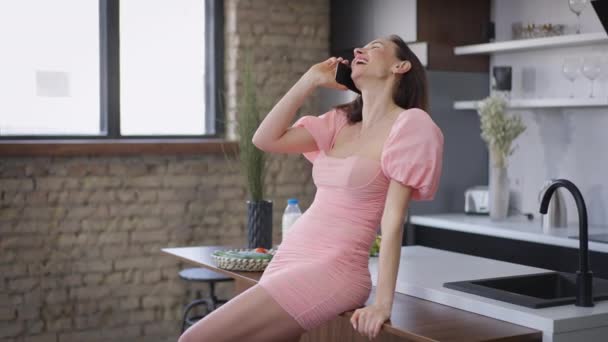 Riéndose hermosa mujer caucásica en vestido rosa hablando por teléfono de pie en la cocina en casa. Alegre relajado hermosa dama confiada en el interior. Movimiento lento. — Vídeo de stock