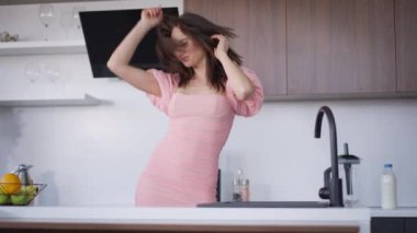 Neşeli, mutlu, rahat, Kafkasyalı, genç, esmer kadın mutfakta ağır çekimde dans ederken eğleniyor. Pembe elbiseli, neşeli, ince bayanın portresi. Hafta sonunun tadını içeride çıkarıyor..