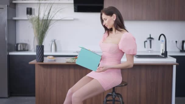 Confiant jeune femme brune en belle robe rose tablette de fermeture et regardant la caméra souriant assis sur la chaise au comptoir dans la cuisine. Portrait de dame mince et charmante posant à la maison à l'intérieur. — Video