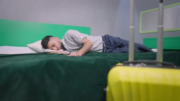 Ευρεία βολή του νεαρού όμορφος άντρας κοιμάται σε άνετο άνετο κρεβάτι με συσκευασμένο κίτρινο ταξιδιωτικό σάκο μπροστά. Μπρουνέτ Καυκάσιος τουρίστας σε εσωτερικούς χώρους με αποσκευές. Αργή κίνηση. — Αρχείο Βίντεο