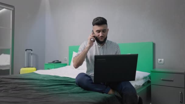 Yakışıklı, başarılı, çok çalışan adamın portresi telefonda konuşuyor ve laptopla yatağa kapanıyor. Kafkas iş adamı yorgun yatak odasında ya da otel odasında proje analizi yapıyor.. — Stok video