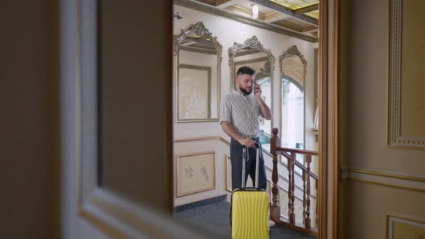 Wide shot reflection in mirror of luxurious hotel of confident young man talking on the phone standing with yellow travel bag in hallway. Portrait of Caucasian tourist indoors with luggage. — Stock Video