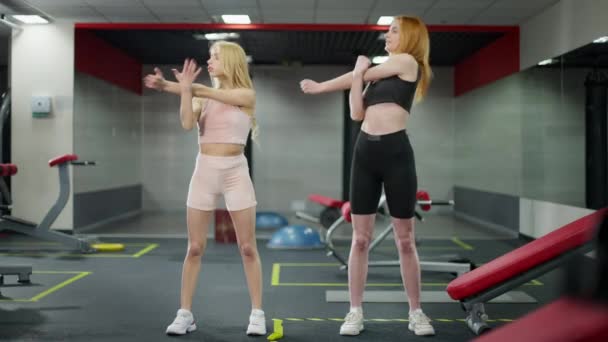 Weitwinkelporträt zweier selbstbewusster junger kaukasischer Frauen, die im Fitnessstudio Hände strecken. Vorderansicht von rothaarigen und blonden Damen beim Aufwärmen im Haus. Gesunder Lebensstil und Sportkonzept. — Stockvideo
