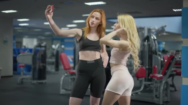 Νεαρές αδύνατες λευκές χιλιετίες γυναίκες βγάζουν selfie στο γυμναστήριο χαμογελώντας και ποζάροντας. Πορτρέτο χαρούμενα όμορφη κοκκινομάλλα και ξανθά χιλιετίες φωτογραφίζοντας για τα μέσα κοινωνικής δικτύωσης. Αθλητισμός και εθισμός στο διαδίκτυο — Αρχείο Βίντεο