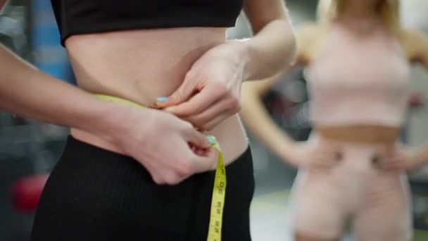 Mujer deportiva irreconocible que mide la cintura en el gimnasio con un entrenador personal de fitness borroso de pie en segundo plano. Resultado después del entrenamiento. Concepto de pérdida de peso y feminidad. — Vídeos de Stock