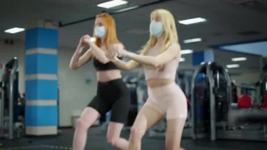 Canlı kamera sarışın ve kızıl saçlı zayıf, beyaz saçlı kadınları takip ediyor. Spor salonunda Covid-19 yüz maskeleri takıyorlar. İki çekici genç sporcu kadın içeride hamleler yapıyor. Coronavirus spor konsepti.