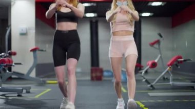 Coronavirus maskeli iki sporcunun spor salonunda hamleler yapmasının ön görüntüsü. Covid-19 salgınında kapalı alanda antrenman yapan kendine güvenen beyaz güzel kadınların portresi. Yeni normal.