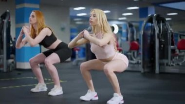Canlı kamera iki sporcuyu spor salonunda çömelirken izliyor. Zayıf, sarışın ve kızıl saçlı kadınların kapalı alanda antrenman yaptığı geniş açılı bir çekim. Spor konsepti.